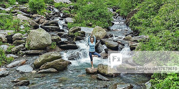 Eine Frau übt die Balance-Yoga-Asana Vrikshasana (Baumhaltung) an einem Wasserfall im Freien. Himachal Pradesh  Indien. Panorama
