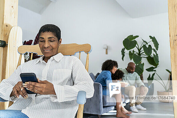 Mature woman sitting on rolling chair with smart phone while family in background
