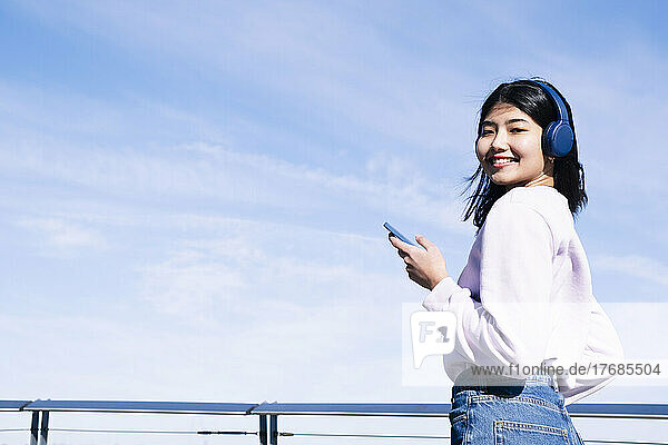 Smiling woman with headphones and smart phone by railing
