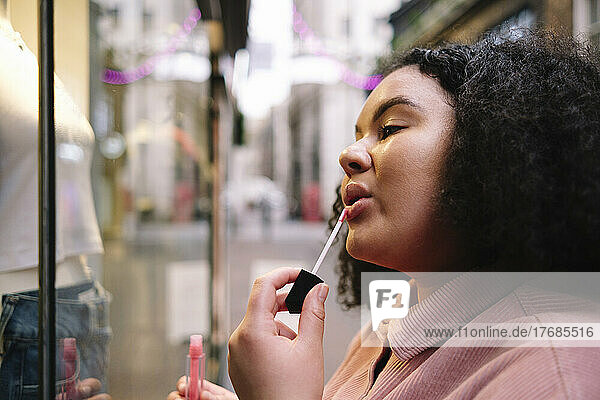 Young woman with curly hair applying lip gloss in front of store window