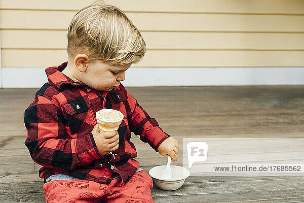 Boy holding ice cream cone with spoon in bowl sitting on porch