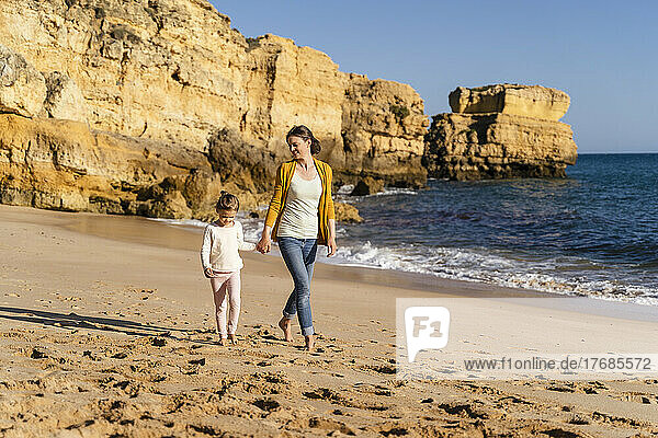 Mother and daughter walking in front of rock formation on shore at beach