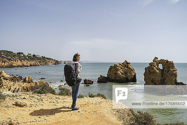 Woman with backpack looking at view standing in front of sea on sunny day