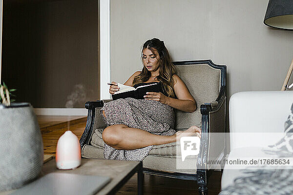 Young woman reading book sitting in armchair at home