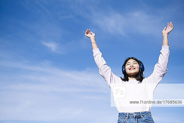 Happy woman listening music with hands raised in front of sky