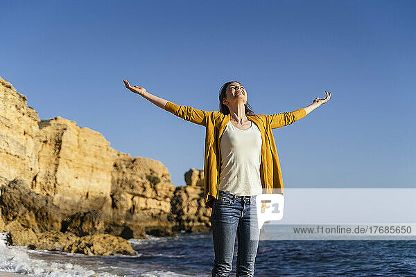 Smiling woman with arms outstretched standing at beach on sunny day
