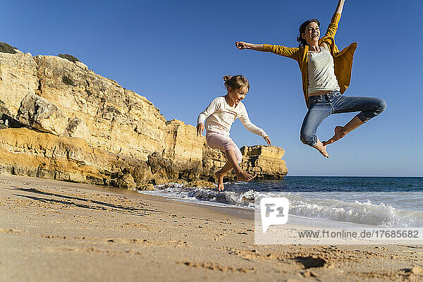 Happy mother with daughter jumping at beach on sunny day