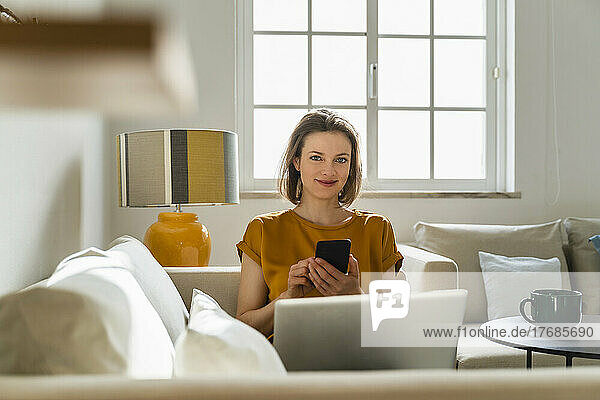 Smiling freelancer with smart phone and laptop sitting on sofa in living room