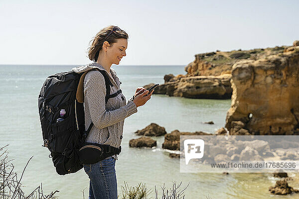 Lächelnde Frau mit Rucksack und Smartphone  die vor dem Meer steht