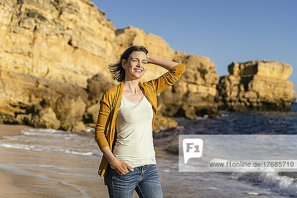 Smiling woman with hand in hair standing at beach on sunny day