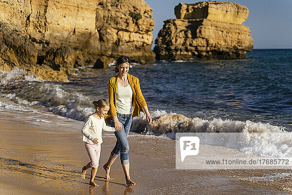 Smiling woman holding hands with daughter walking on shore at beach