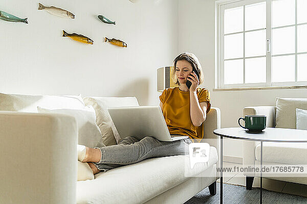 Freelancer talking on smart phone using laptop sitting on sofa at home