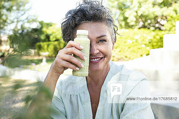 Smiling woman covering face with bottle of smoothie sitting in garden