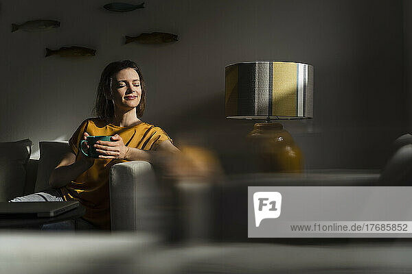 Smiling woman with eyes closed holding coffee mug sitting in living room
