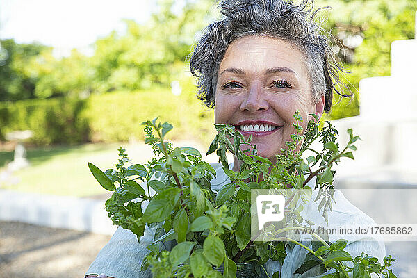 Lächelnde reife Frau mit Pflanze sitzt an einem sonnigen Tag im Garten