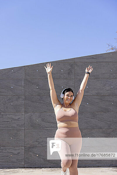 Smiling woman listening music through wireless headphones exercising with hands raised in front of wall