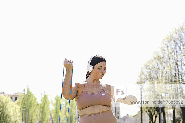 Smiling woman listening music through wireless headphones and exercising with resistance band in park