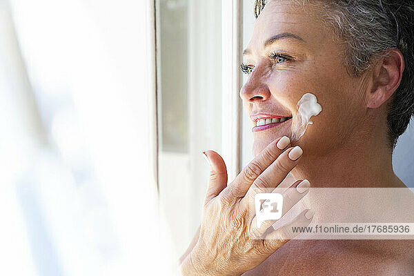 Smiling mature woman applying moisturizer cream on face