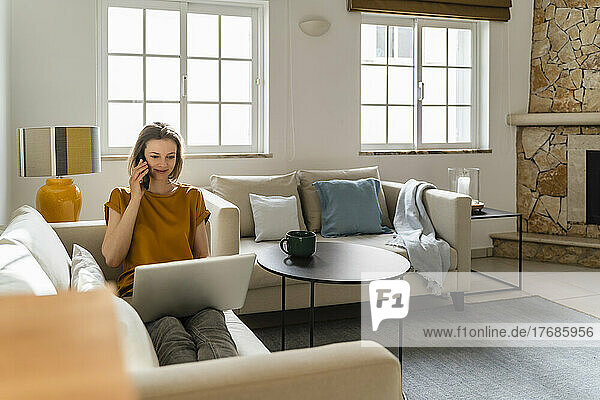 Smiling freelancer talking on smart phone sitting with laptop working at home