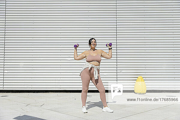 Smiling curvy woman holding dumbbells standing in front of corrugated wall