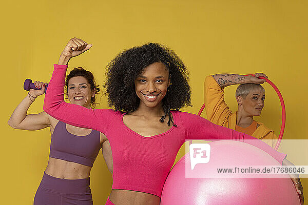 Multiracial friends with fitness equipment standing against yellow background