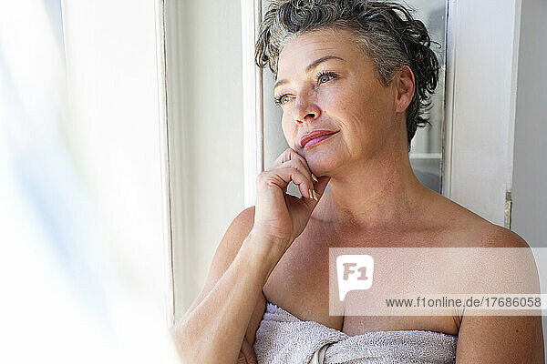 Thoughtful woman wearing towel standing with hand on chin at home
