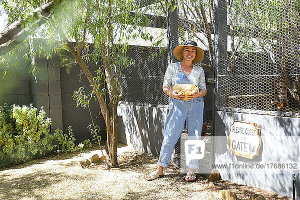 Smiling farm owner wearing hat holding bowl at entrance on sunny day