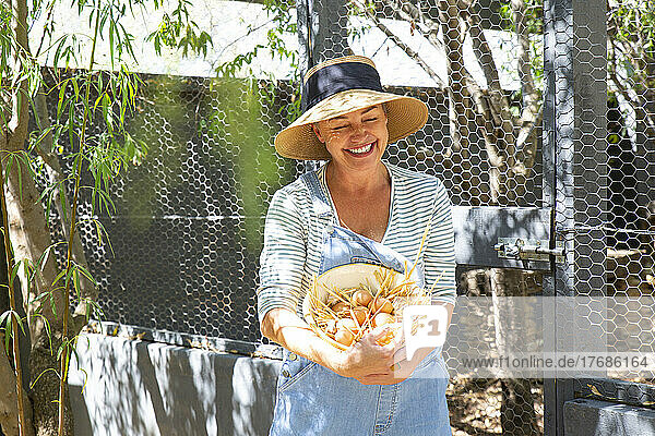 Happy mature farm owner holding egg's bowl in front gate on sunny day