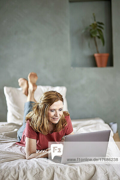 Smiling woman using laptop lying on bed at home