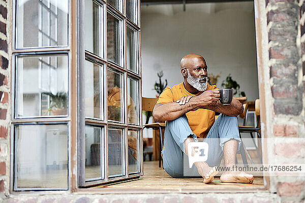 Thoughtful bald man with coffee mug sitting by window