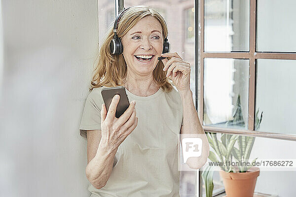 Glückliche Frau mit Headset und Smartphone vor dem Fenster zu Hause