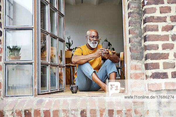 Bald man text messaging through smart phone sitting by window
