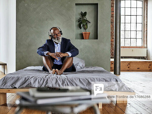 Businessman listening music through wireless headphones sitting on bed at home