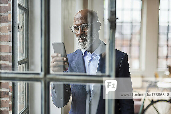 Businessman with smart phone seen through window at home