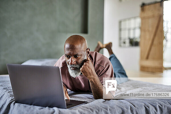 Mature man with white beard using laptop lying on bed at home