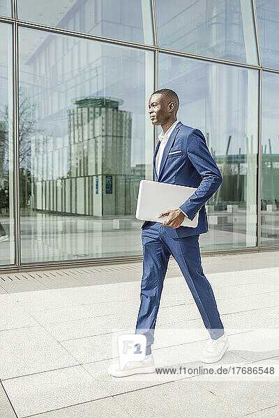 Businessman holding laptop walking by modern office building