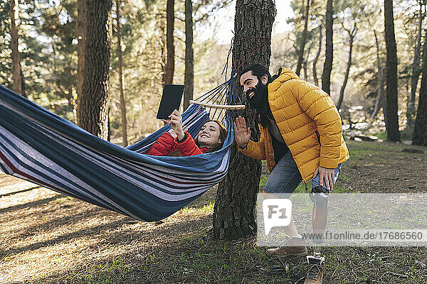 Smiling disabled man waving by woman doing video call through tablet PC lying in hammock