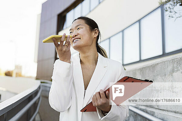 Happy businesswoman holding file folder talking on mobile phone through speaker