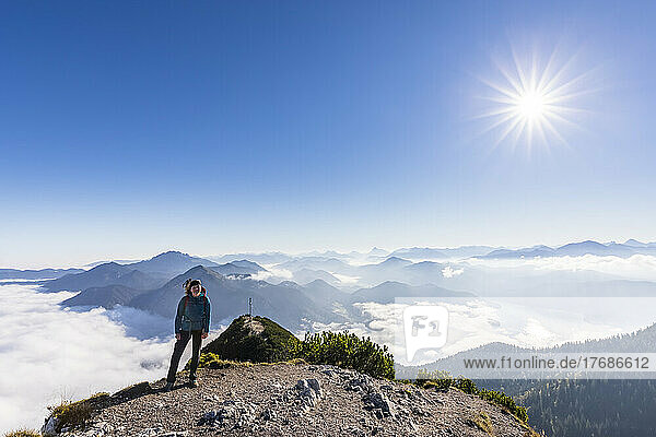 Germany  Bavaria  Sun shining over female hiker standing on mountaintop with thick fog and peak of Herzogstand mountain in background
