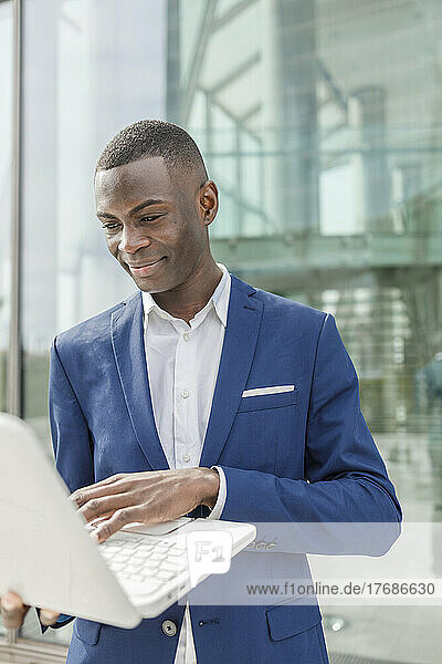 Smiling businessman using laptop in front of modern office building