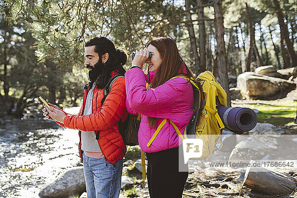 Man using smart phone by girlfriend looking through binoculars in forest