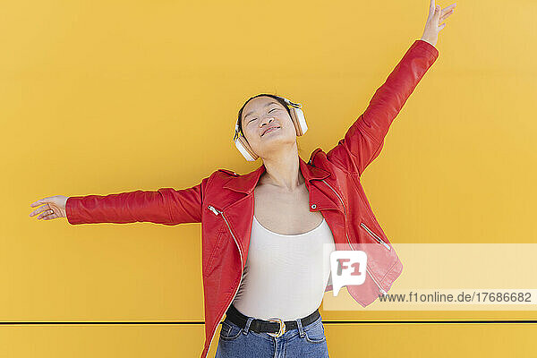 Young woman listening music through wireless headphones in front of yellow wall