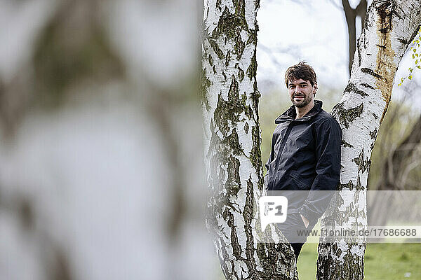 Smiling man with hand in pocket leaning on tree trunk