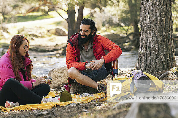 Smiling bearded man looking at girlfriend sitting on picnic blanket in forest