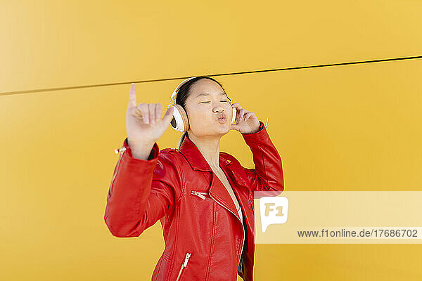 Junge Frau hört Musik über drahtlose Kopfhörer und zeigt Shaka-Schild vor gelber Wand