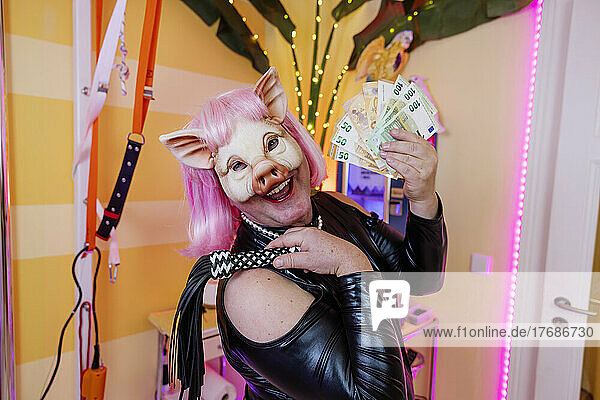 Happy transvestite man wearing pig mask holding whip and paper currency