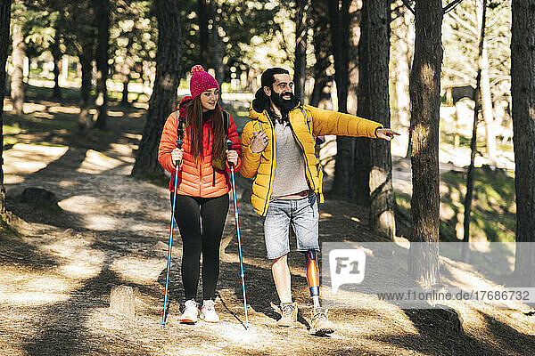 Happy disabled man pointing away by woman walking with hiking pole in forest