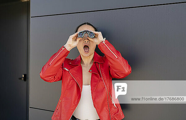 Schockierte Frau mit offenem Mund blickt durch ein Fernglas vor einer grauen Wand
