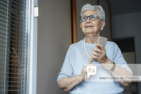 Happy senior woman with smart phone standing at apartment doorway