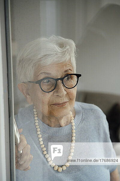 Curious senior woman wearing eyeglasses looking through glass window at home